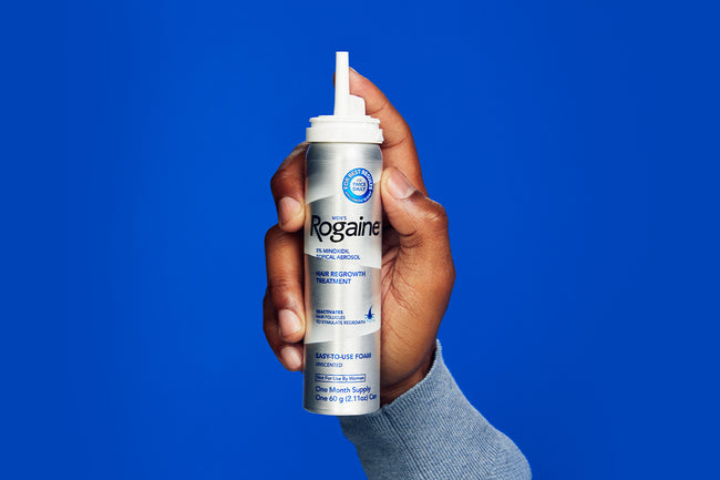 Close-up image of a hand holding Men's ROGAINE® Foam Solution on a blue background.