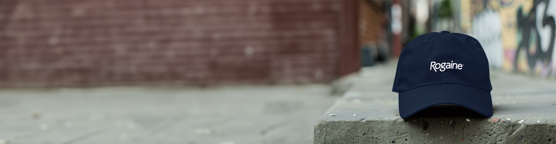 Navy blue baseball cap with a ROGAINE® logo, resting on top of a bench, with a brick building in the background.