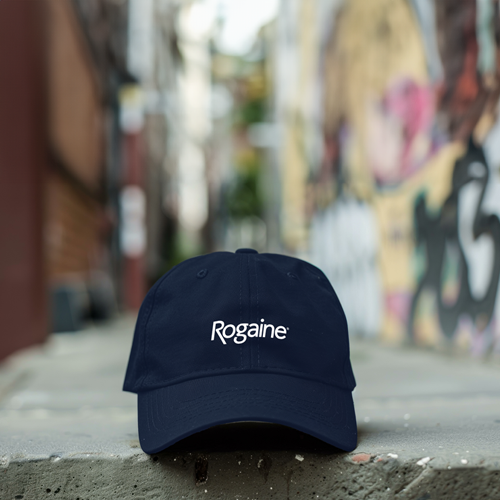 Navy blue baseball cap with a ROGAINE® logo, resting on top of a bench, with a brick building in the background.