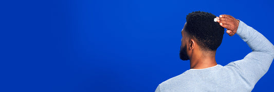Man with dark brown hair and beard applying Men's ROGAINE® Foam to the back of his head on a blue background.