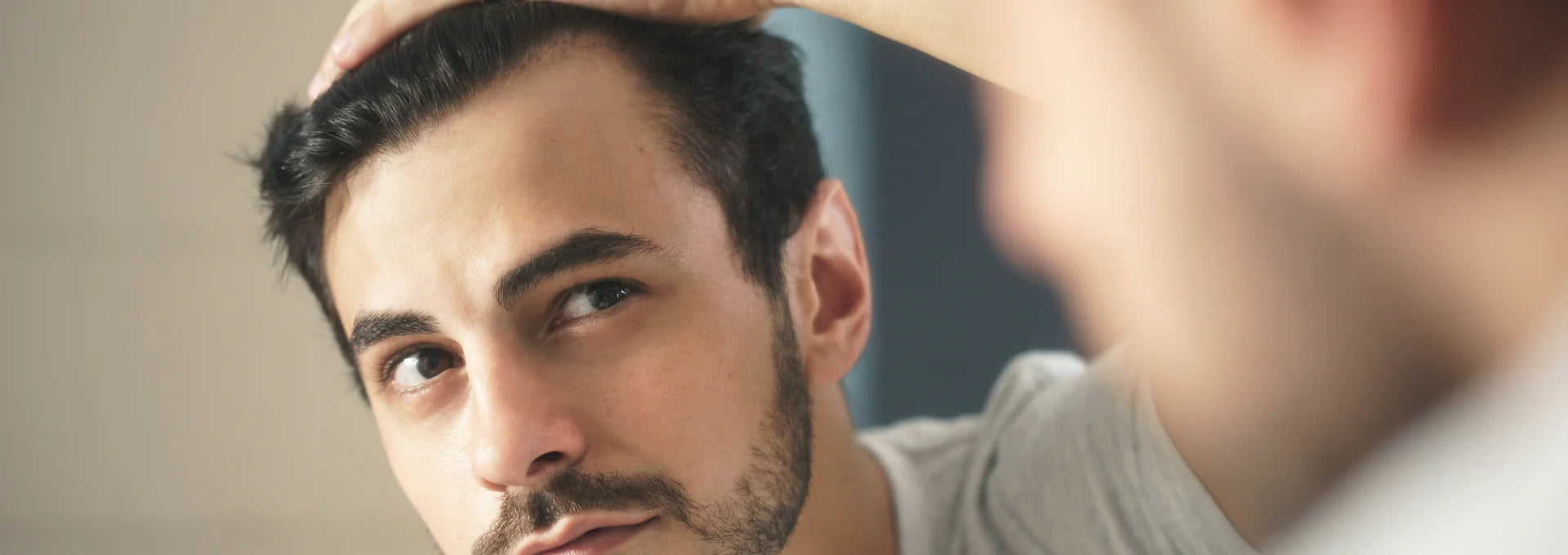 Man with dark brown hair and beard checking his hairline in the mirror.