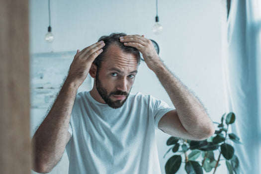 A man with light brown hair and a beard inspecting the part of his hair in a mirror. 