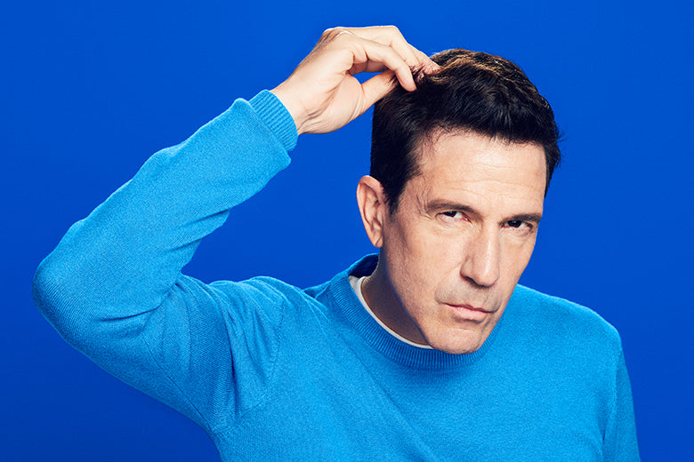 Man with brown hair and a long sleeve blue shirt touching his hair, on a blue background.