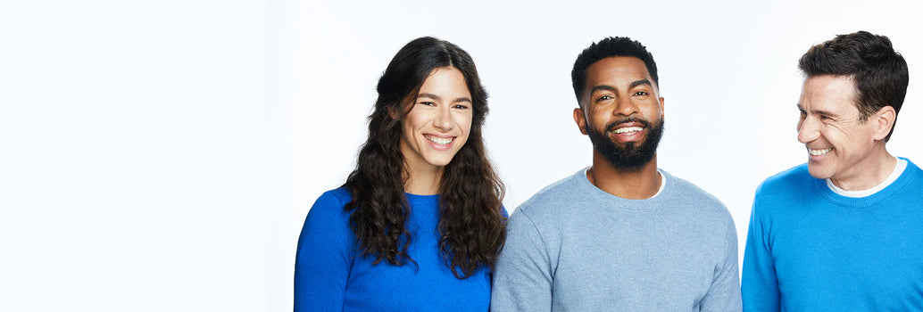 Woman with long brown hair smiling next to two men with dark hair wearing long sleeve shirts on a white background.